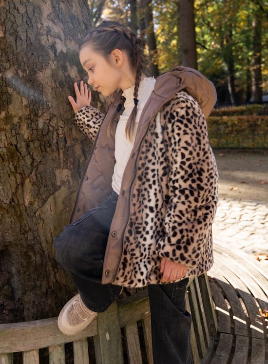 Meisje met vlechten draagt bruine jas met leopardprint, leunt tegen boom.