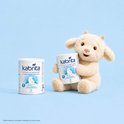 Een blik Kabrita zuigelingenmelk met geitenmelk en een knuffelgeitje, op een blauwe achtergrond.