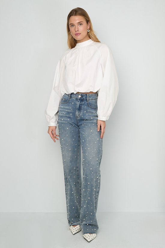 Model in een spijkerbroek met pareltjes en een witte blouse met hoge hals en lange mouwen.