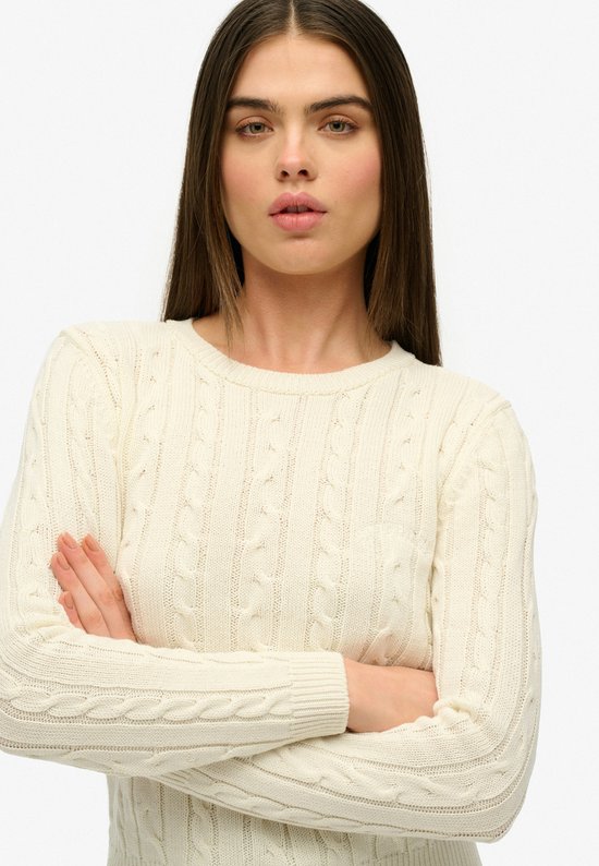 Gros plan sur un mannequin dans un pull torsadé Superdry de couleur crème, bras croisés.