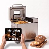RVS broodbakmachine met open deksel, waarin zaden op vers brood vallen. Een tablet met "Met uitleg Video's" wordt ernaast vastgehouden.