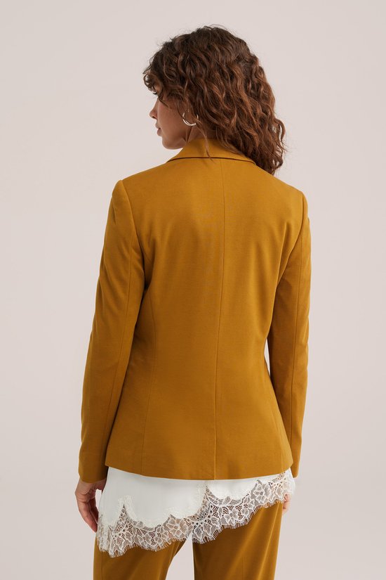 Achterkant van okergele jersey blazer, gedragen door model met witte top.