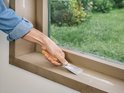Hand gebruikt een plamuurmes om witte vulmiddel uit te strijken op een houten vensterbank naast een raam.