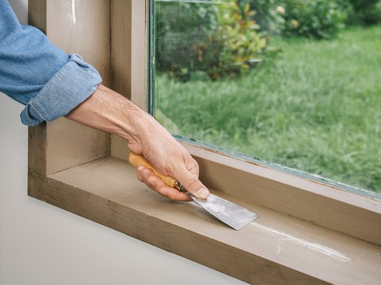 Hand gebruikt een plamuurmes om witte vulmiddel uit te strijken op een houten vensterbank naast een raam.