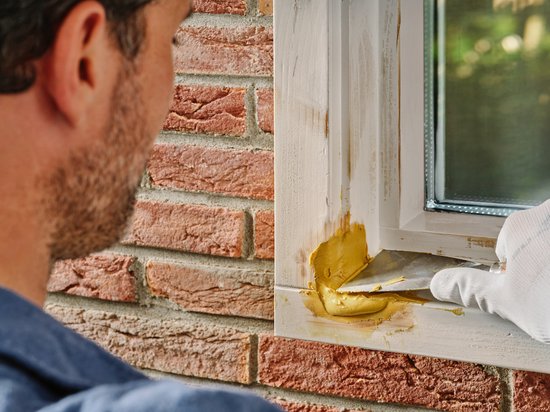 Man met grijze baard en witte handschoen schuurt een hersteld wit kozijn.