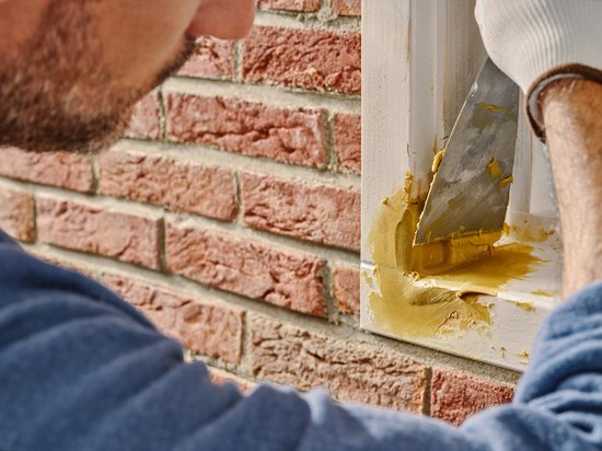 Man met witte handschoen smeert gele houtrotvuller op een beschadigd wit kozijn met een spatel.