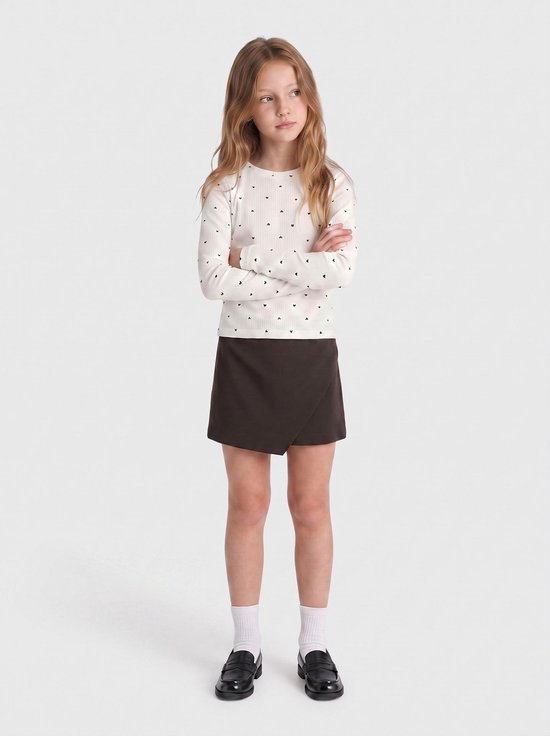 Wit shirt met lange mouwen en zwart hartjespatroon, gedragen door een meisje met een bruine omslagrok, witte sokken en zwarte schoenen.