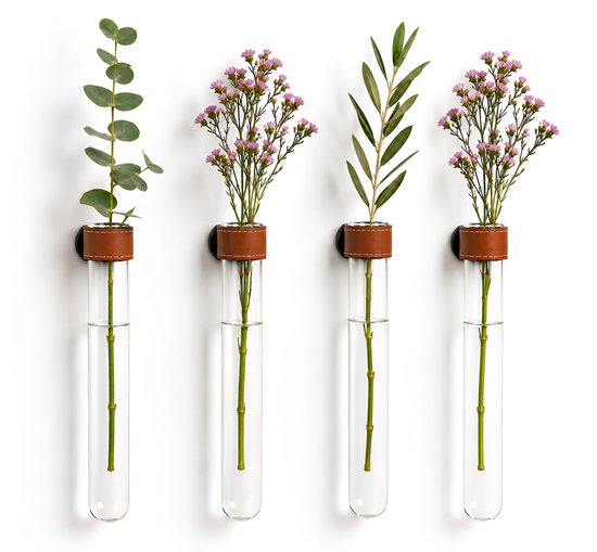 Vier glazen reageerbuisjes met takken, bloemen en bladeren, elk met een bruin leren bandje en zwarte magneet, opgehangen aan een witte muur.