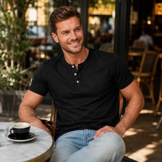 Man met bruin haar, lacht, draagt zwart henley shirt en lichtblauwe jeans, zit buiten bij een tafel.