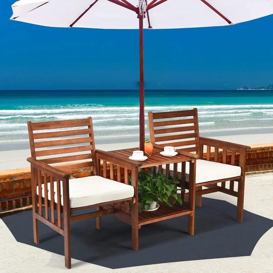 Een bruine houten tuinset met witte kussens en een tafel met parasol, op een terras aan zee. De tafel heeft een plant en twee kopjes.