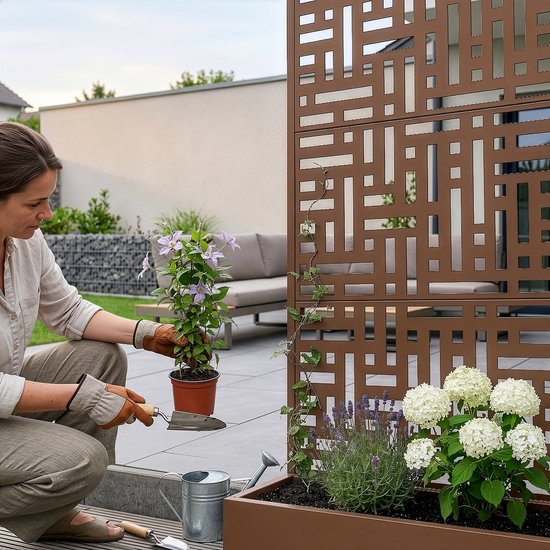 Een vrouw plant een klimplant in een bruine plantenbak naast een tuinscherm met een geometrisch patroon.