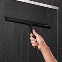 Une main utilise un racloir de douche noir pour enlever l'eau d'une paroi de douche carrelée de couleur foncée.