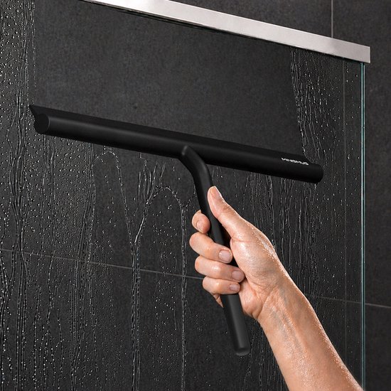 Une main utilise un racloir de douche noir pour enlever l'eau d'une paroi de douche carrelée de couleur foncée.