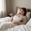 Een lachende vrouw met een beige trui ligt in bed met een crème warmtekussen in haar handen. Het kussen heeft een zwart label.
