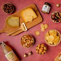 Borrelplank met kazen, worstjes, crackers, chips, noten, dip en sap. Alles op een rode achtergrond.