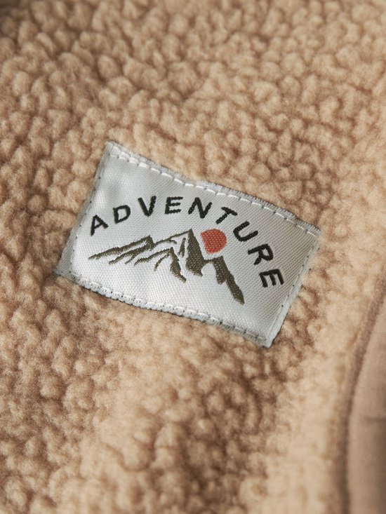 Close-up van een 'Adventure' patch op een beige teddy stof. De patch toont bergen en de tekst 'Adventure'.