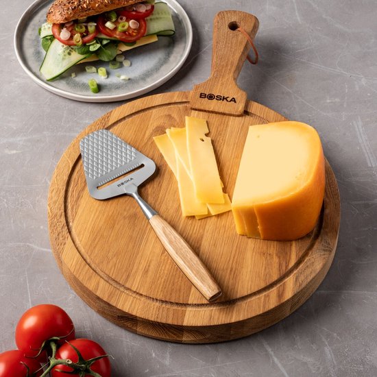 Houten kaasplank met een stuk kaas, plakjes kaas en een kaasrasp met houten handvat. Tros tomaten en broodje op de achtergrond.