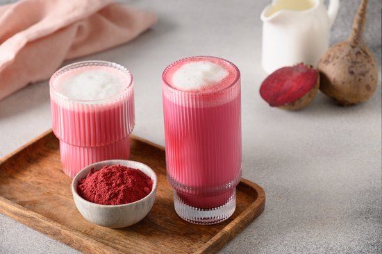 Twee glazen roze bietendrank, met schuim, op een houten plank met een bakje bietepoeder en biet.