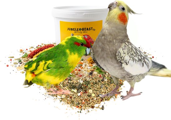 Kakariki met groene borst en rode kop, en een valkparkiet met grijze veren en gele kuif, zittend op vogelzaad met een Jungle Beast emmer.
