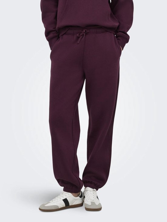 Model draagt een bordeauxrode joggingbroek met koord en witte sneakers met zwarte strepen.