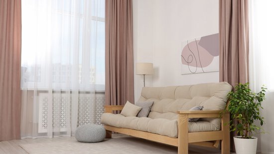 Woonkamer met beige bank, grijze poef en taupe gordijnen voor een raam.