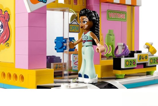 Figurine aux cheveux tressés et taches de naissance à l'entrée d'un magasin rose et jaune avec un panneau 'OUVERT !'.