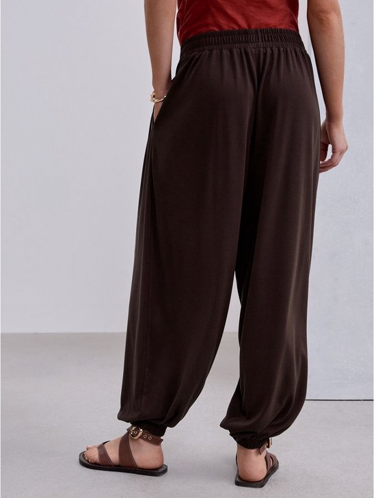Achterkant van een model in bruine ballonbroek en roodbruine top, met bruine sandalen.