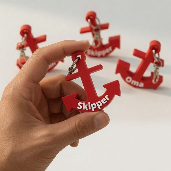 Hand houdt rode 'Skipper' ankersleutelhanger vast. Drie andere rode ankers zijn op de achtergrond.