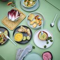 Een tafel met een zomerse maaltijd: salades, brood, dips en fruit. Diverse lichtgroene en grijze schalen staan verspreid.