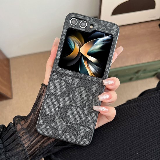 Een Samsung Z Flip 5 telefoonhoesje met grijs-pu leer en Coach-logo patroon, vastgehouden door een hand met lange nagels.