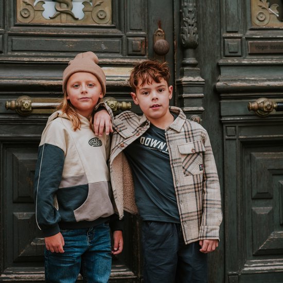 Twee jongens die voor een donkergroene deur staan. De jongen links draagt een beige en grijze hoodie. De jongen rechts draagt een geruit jasje met teddy voering over een donkergrijs shirt.