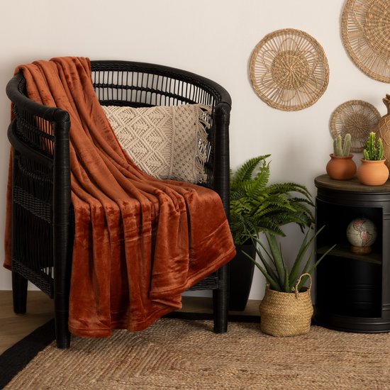 Plaid en polaire couleur terre cuite, 130 x 180 cm, sur un fauteuil noir avec coussins et plantes.