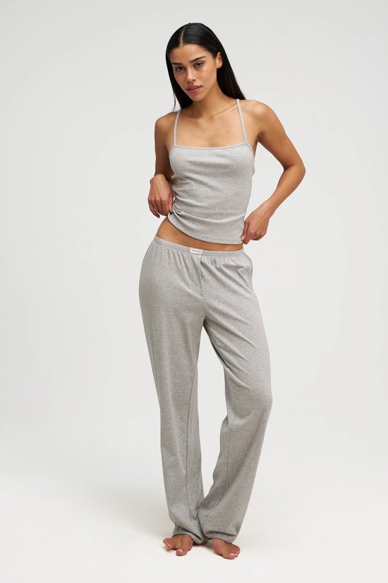 Model in een grijze pyjama met spaghettibandjes top en wijde broek, staand op witte achtergrond.