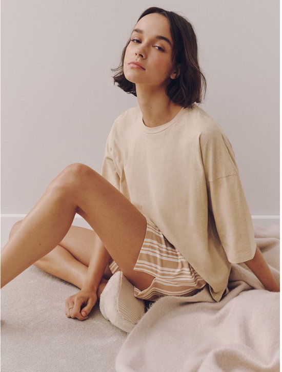 Model zittend op de vloer in een beige pyjamaset, met effen top en gestreepte short.