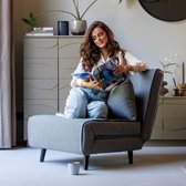 Een vrouw met lang bruin haar, gekleed in een witte blouse en jeans, zit ontspannen in een comfortabele, grijze slaapstoel en leest een tijdschrift.