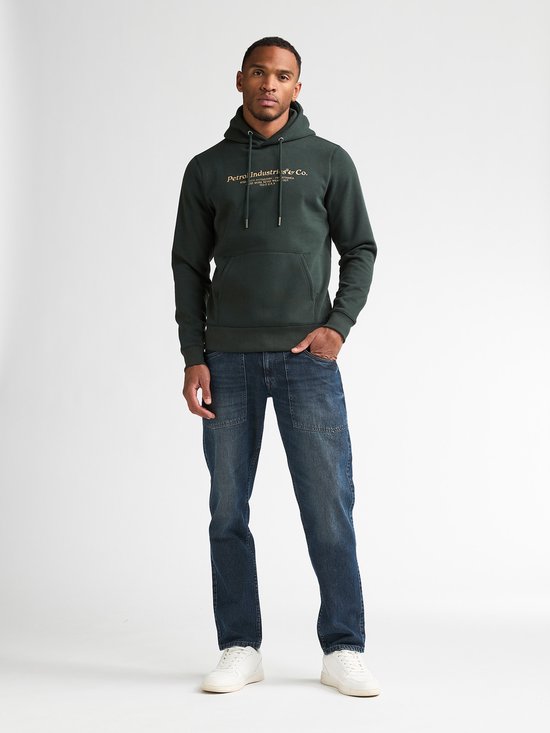 Man draagt een donkergroene hoodie met gouden tekstprint op de borst en spijkerbroek.