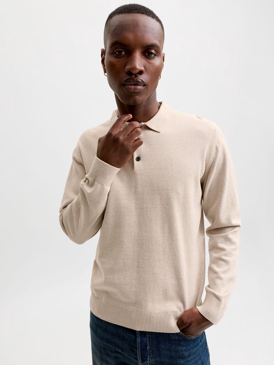 Man, lichtbeige gebreid poloshirt met lange mouwen, twee zwarte knoopjes en donkerblauwe jeans.