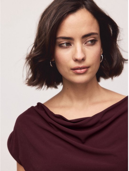 Close-up van een vrouw met korte bruine haren en zilveren oorbellen, die een diepbruine top met gedrapeerde hals draagt.