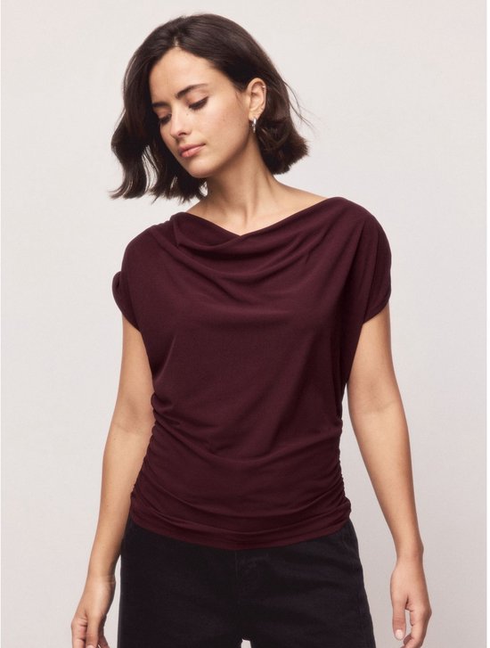 Een vrouw met korte bruine haren draagt een diepbruine mouwloze top met gedrapeerde hals en zwarte broek.