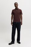 Man draagt bordeauxrode polo met witte en marineblauwe streepdetails, met donkere jeans.