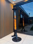 Zwarte pelletkachel met vlammen staat op een terras naast een modern gebouw met glazen pui.