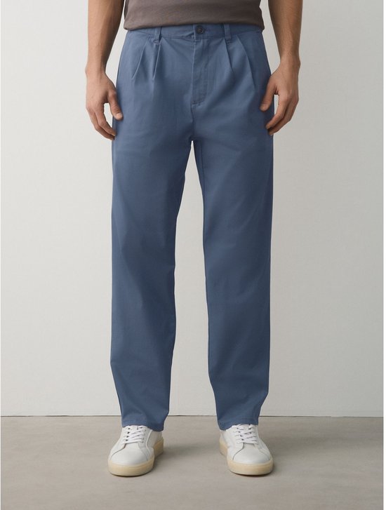 Vooraanzicht van een man in een losse, blauwe chino met plooien en witte sneakers.