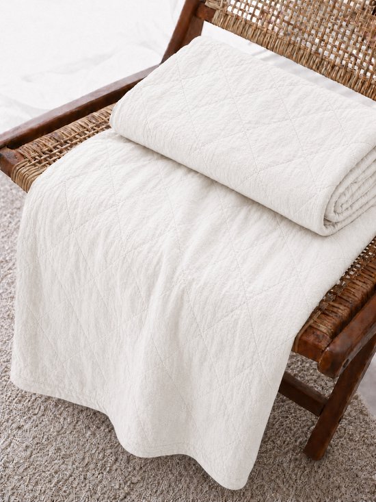 Witte, gewatteerde sprei met ruitpatroon, opgevouwen op een houten stoel.