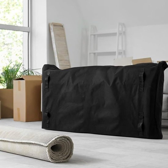 Gesloten zwarte matras beschermhoes met handvatten, staand op een vloer met verhuisdozen en een opgerolde deken.