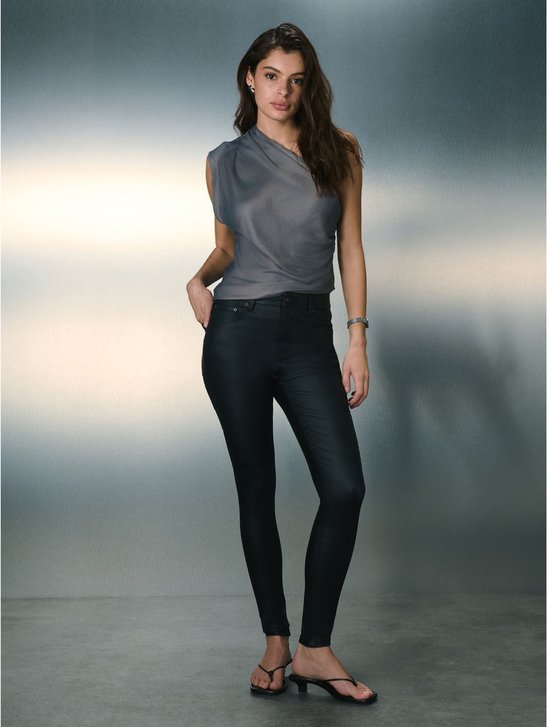Model staand, gekleed in een zwarte skinny coated broek en grijze top. Ze draagt platte zwarte sandalen.