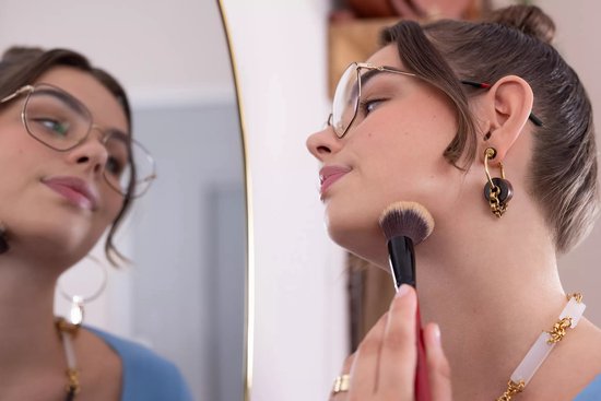 Jonge vrouw met bril gebruikt een make-up kwast op haar kaaklijn, in het spiegelend oppervlak van een spiegel.