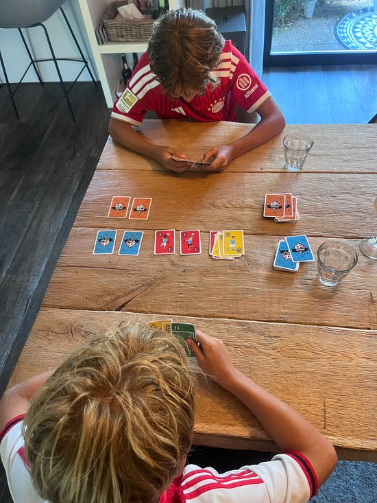 Twee jongens met rode en witte voetbalshirts spelen kaartspel aan een houten tafel. Kaarten met voetballers.