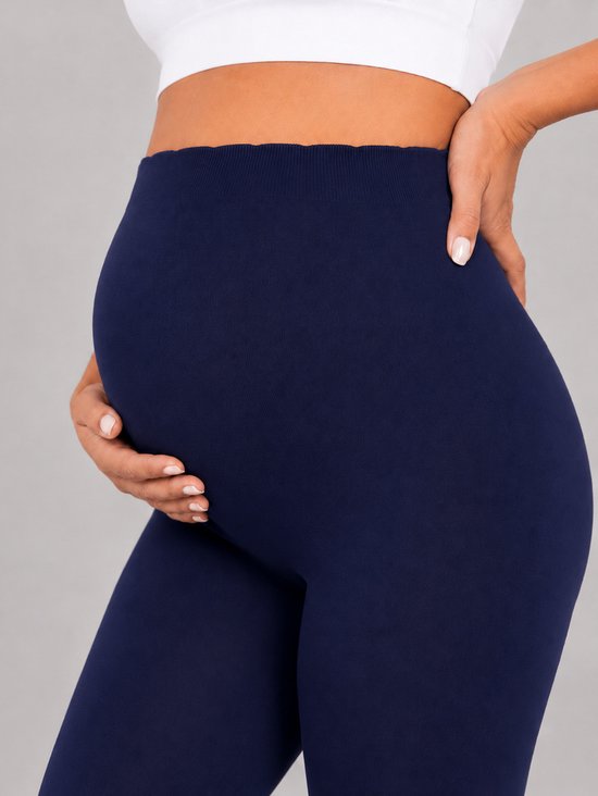 Zij-aanzicht van een zwangere vrouw die haar buik vasthoudt, draagt een donkerblauwe legging.