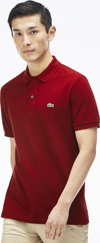 Een man draagt een rode Lacoste polo met korte mouwen en het groene krokodillenlogo, gecombineerd met een beige broek.