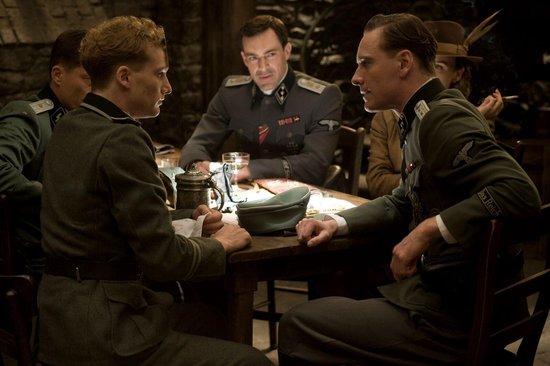 Vier mannen in Duitse uniformen zitten aan een tafel, met twee spelkaarten op hun voorhoofd, in een gedempt verlichte ruimte.
